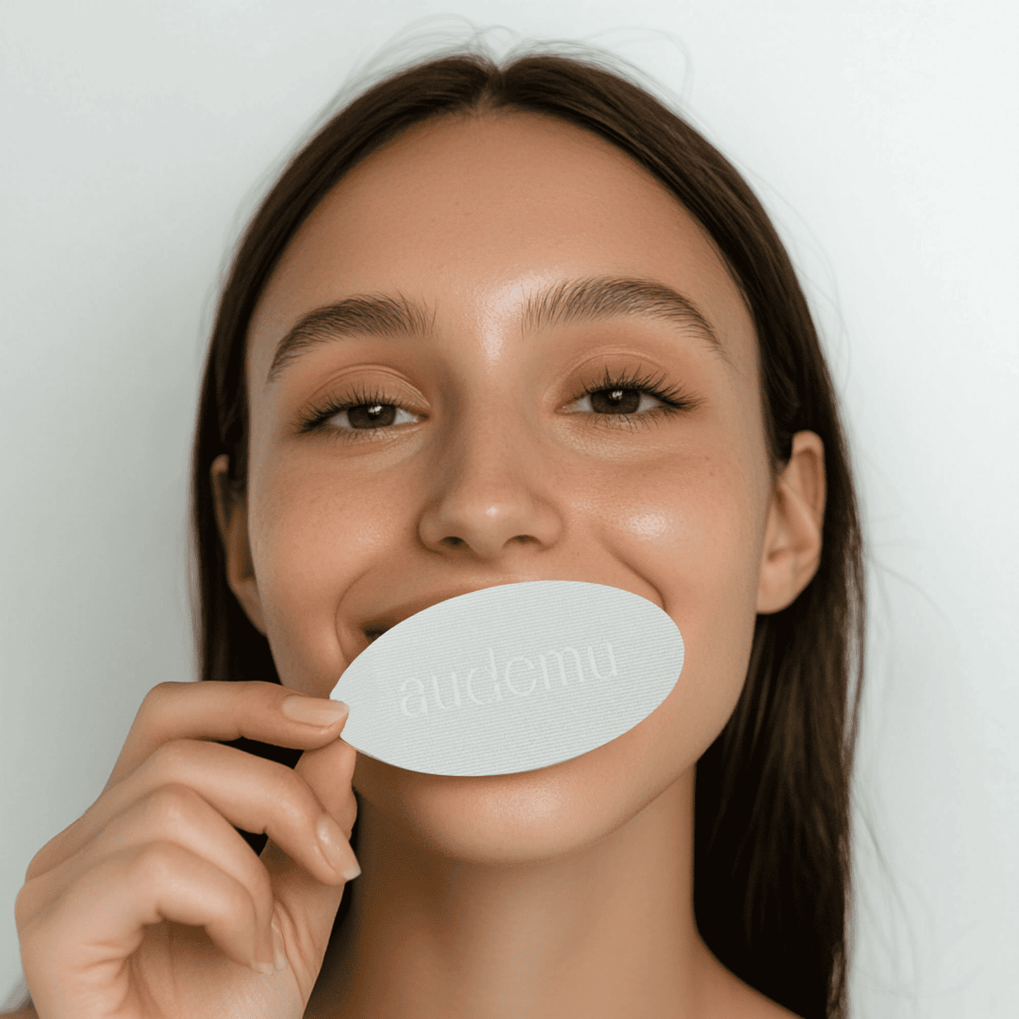 Frau mit langen dunklen Haaren hält ein weißes ovales Audemu-Produkt zwischen den Zähnen – lächelnd vor weißem Hintergrund. Visuelle Darstellung der Marke und des Produkts für Beauty-Routinen oder nächtliche Pflege.
