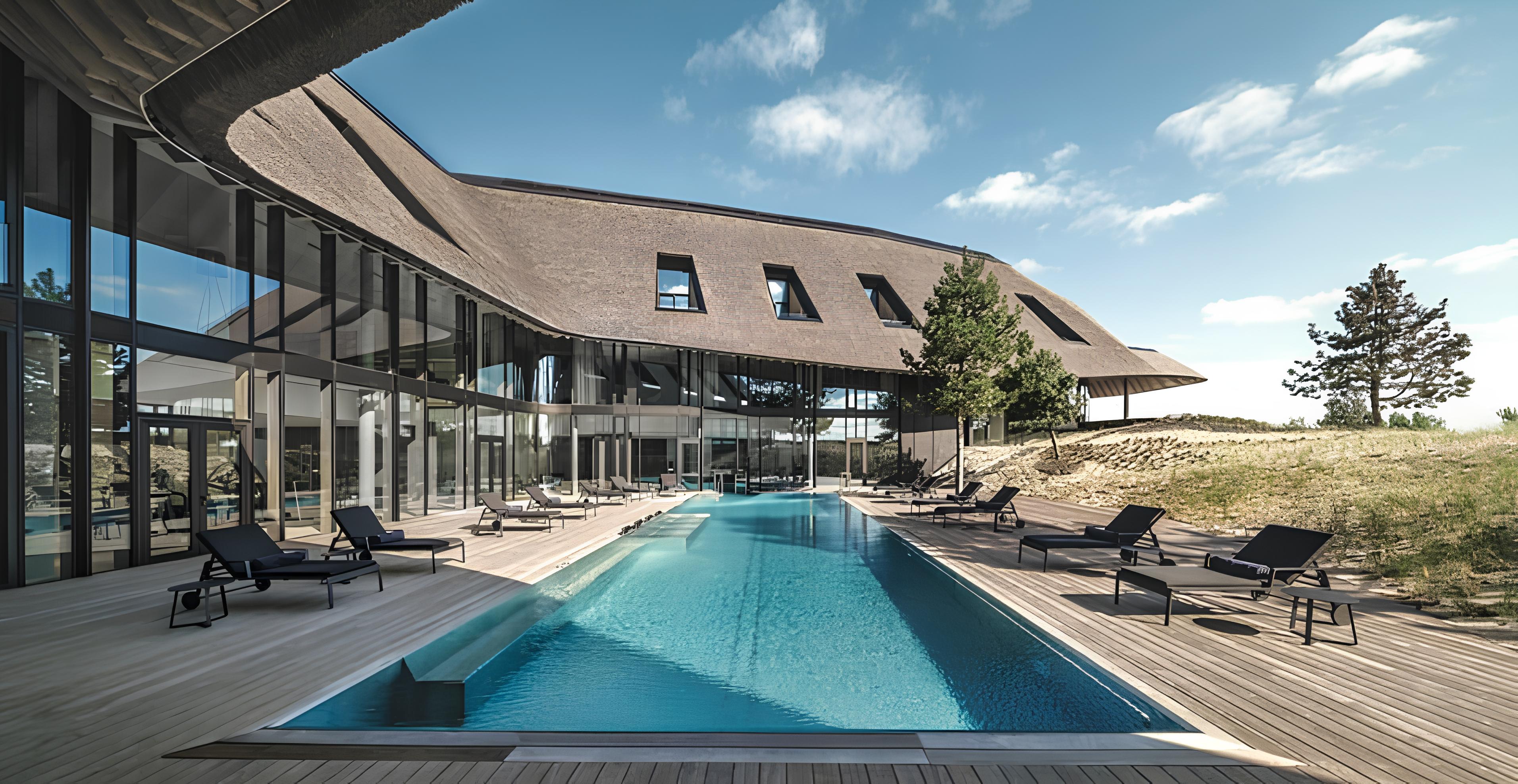 Modernes Luxus-Spa mit wellförmigem Strohdach und Glasfront – langer Pool mit hellblauem Wasser auf Holzterrasse mit Liegestühlen. Umgeben von natürlicher Landschaft unter blauem Himmel – stilvolle Architektur im Einklang mit der Umwelt. Ein typisches Sylt feeling.