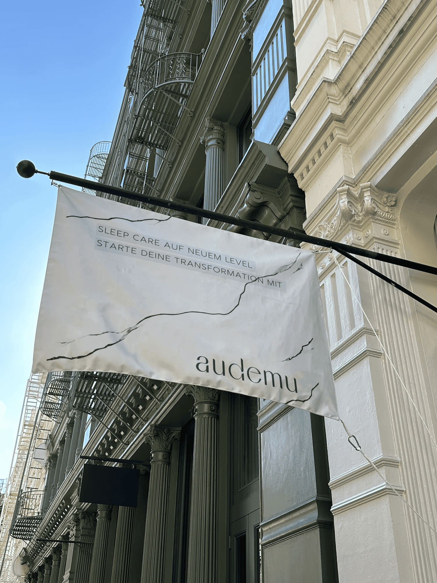 Audemu-Werbefahne hängt an einem historischen Gebäude – Text auf der Fahne: „SLEEP CARE AUF NEUEM LEVEL. STARTE DEINE TRANSFORMATION MIT AUDEMU“. Die Fahne ist weiß mit schwarzem Schriftzug und hängt vor einer klassischen Fassade mit Balkonen und Säulen.