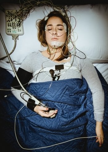 Frau schläft in einem Behandlungszimmer. mit Elektroden am Kopf und Brust zur Überwachung der Schlafqualität – sie trägt ein Gewichtsdecke und ist an einen Schlafmonitor angeschlossen. Ihre Augen sind geschlossen.